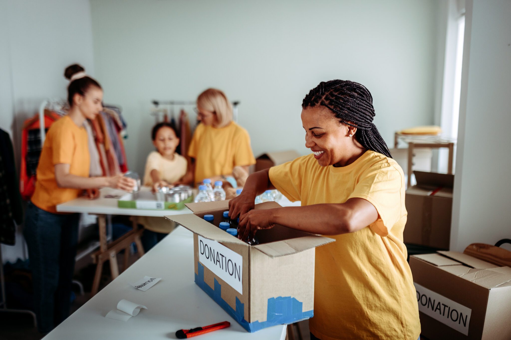 Smiling woman organizing donations in the foreground, while her colleagues collaborate in the background. The scene captures teamwork and dedication in a busy volunteer center. Ideal for themes of community service, charity work, and cooperative effort