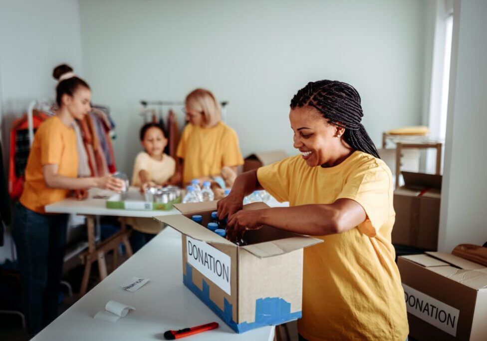 Smiling woman organizing donations in the foreground, while her colleagues collaborate in the background. The scene captures teamwork and dedication in a busy volunteer center. Ideal for themes of community service, charity work, and cooperative effort